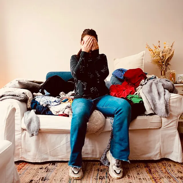 Vrouw zit met de handen voor haar gezicht op een bank vol stapels kleding